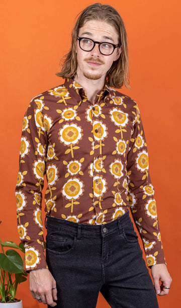 Jack a white male with shoulder length blonde hair and glasses in his 20's wearing a retro brown shirt with long selves and black jeans shot against an orange studio background