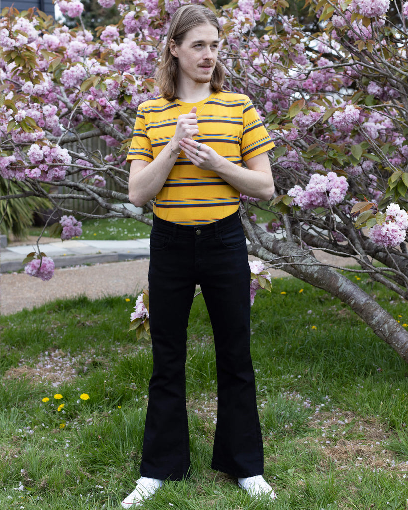 Jack is a tall blonde male with shoulder length hair looking into camera and smiling wearing a yellow retro tee and black bell bottom flares against a pink blossom tree