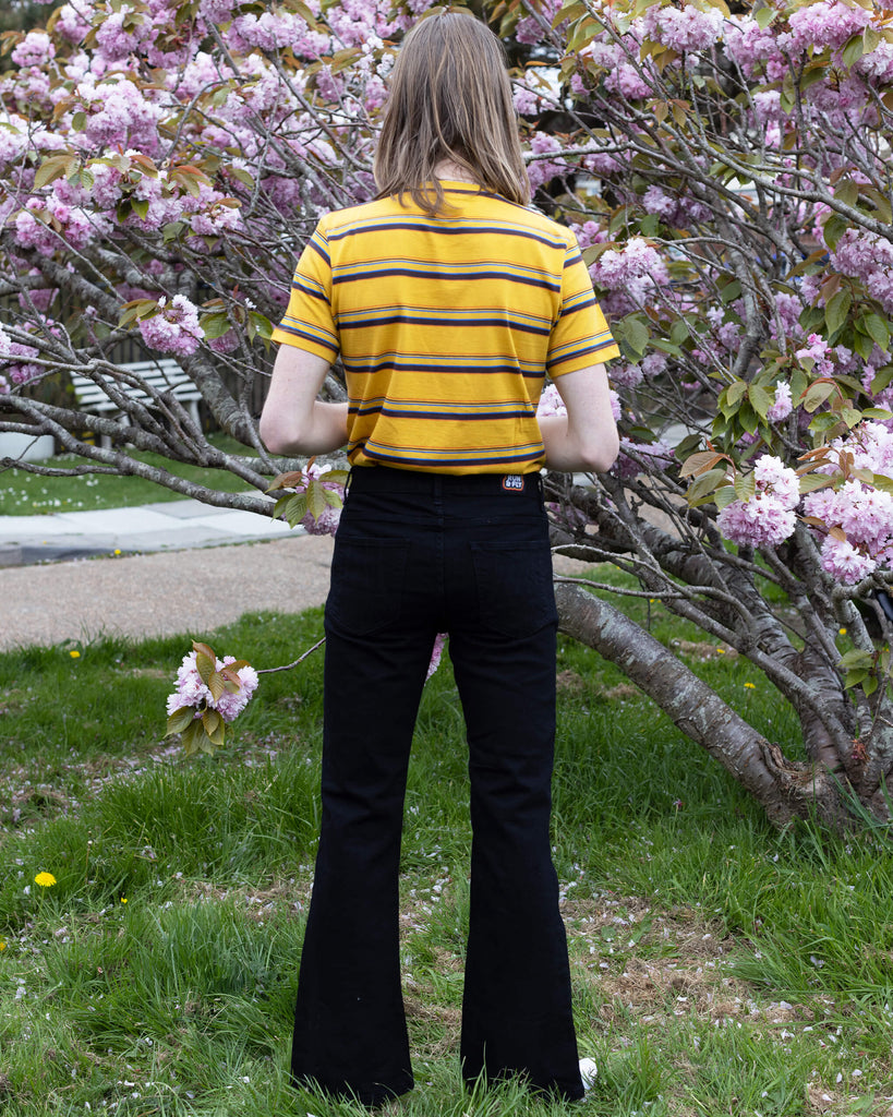 Jack is a tall blonde male with shoulder length hair looking into camera and smiling wearing a yellow retro tee and black bell bottom flares against a pink blossom tree he is facing the back