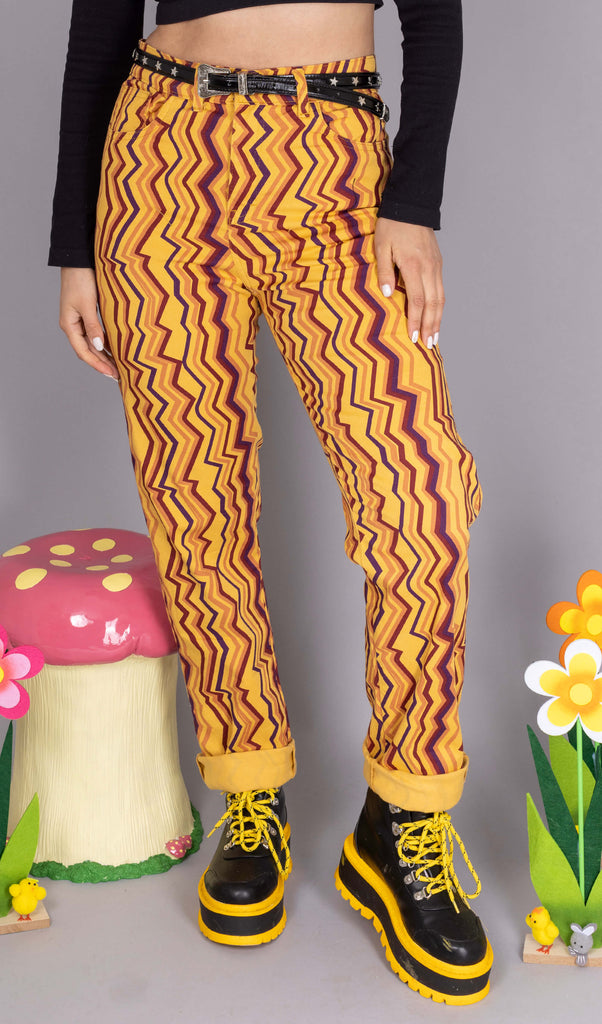 Shannon is stood in front of a grey studio background wearing the 90's zigzag straight leg high waisted jeans with a long sleeve black tshirt and yellow black boots. Surrounding her are floral stand decorations and a mushroom stool. She is facing forward posing with one leg out to the side and both arms resting by her side. Photo is cropped from the waist down.