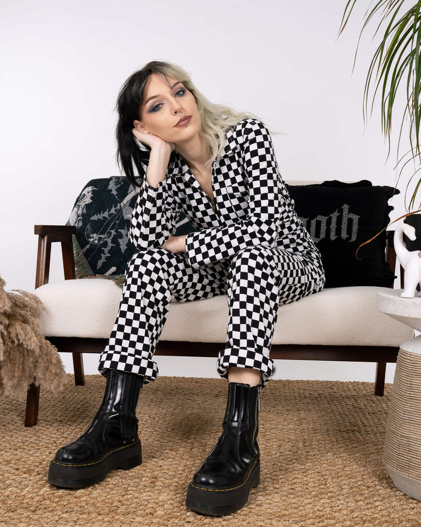 A full-body shot of a model seated on a modern sofa in the Black & White Checkerboard Stretch Twill Boiler Suit by Run & Fly. Styled with chunky black boots, the sustainable and edgy look showcases ethical slow fashion.