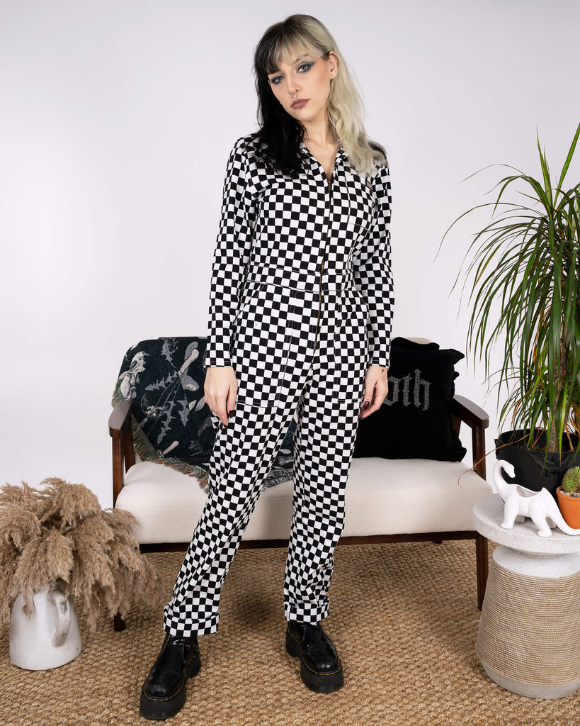 A full-body image of a model wearing the Black & White Checkerboard Stretch Twill Boiler Suit by Run & Fly. Featuring a bold checkerboard print, collared neckline, and gold zip front, paired with chunky black boots. Ethical slow fashion meets edgy statement style, perfect for a sustainable wardrobe. The scene includes natural accents like pampas grass and plants, adding a contemporary vibe.