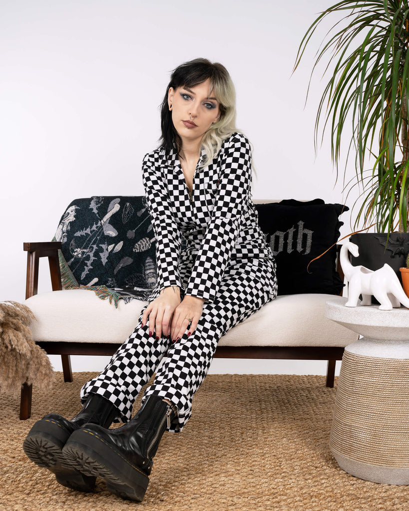 A seated view of the Black & White Checkerboard Stretch Twill Boiler Suit by Run & Fly. The bold pattern contrasts with the model's chunky black boots, epitomising edgy, ethical slow fashion.