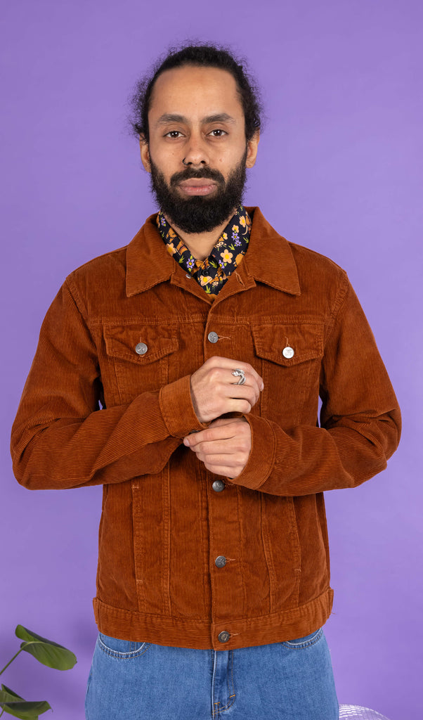 Richard, a hispanic male model with dark hair in a bun and a beard, is stood in a photography studio in Hove in front of a lilac backdrop wearing Retro Vintage Tan Corduroy Unisex Western Jacket buttoned up with Highland Cows Short Sleeve Shirt underneath and blue jeans. Richard is posing to the camera and is doing up the button one of the jacket sleeves. The photo is cropped at the thighs.