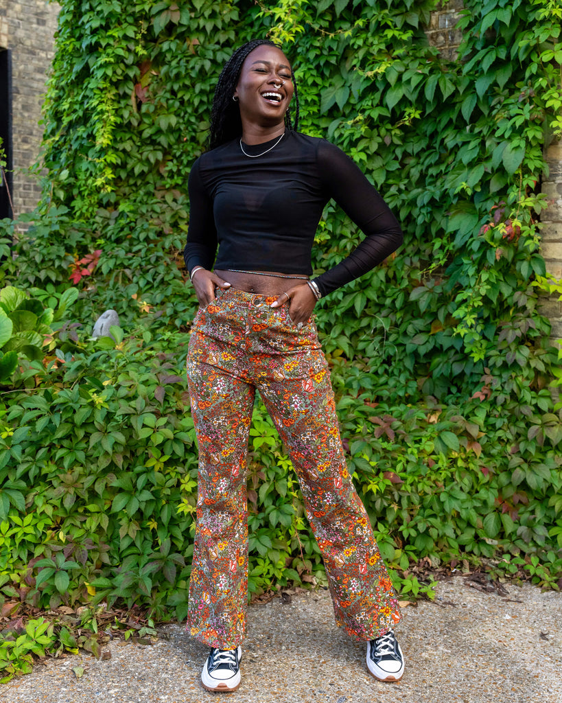 Yolande models a pair of vibrant floral flared trousers from Run & Fly, paired with a black long-sleeve mesh top. She’s laughing with her hands in her pockets, showing off the comfortable fit and playful style. The green backdrop highlights the ethical slow fashion focus of the brand.