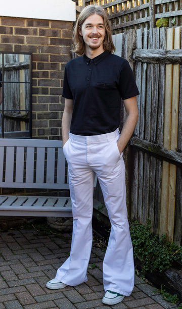 Jack is stood outside in a garden area wearing the Presley white cotton bell bottom trouser slacks with a short sleeve black shirt and white trainers. They have mid length blonde hair and are facing the camera posing with both hands in their front trouser pockets whilst looking off to the right smiling.