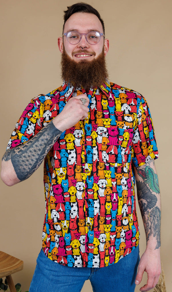 Declan a white bearded tattooed man in his 20's with brown hair and glasses is smiling at camera wearing a multicoloured shirt with dogs on and blue jeans on a beige backdrop shot in studio