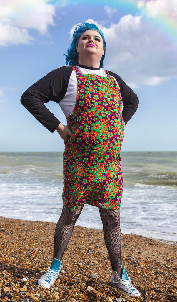 Jeff is stood on a beach wearing the 70s dark floral print stretch corduroy long pinafore dress with a long sleeve black tshirt, tights and white trainers. They are facing forward posing with both hands on their hips and smiling looking up.