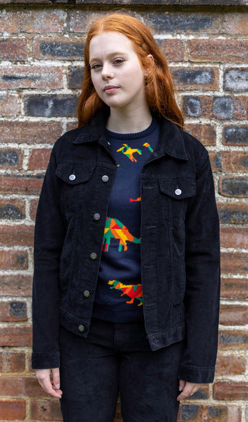 Sophie is stood in front of a brick wall wearing the retro vintage black cord unisex western jacket with the rainbow dino jumper and black trousers. She is facing forward with both arms resting by her sides. Photo is cropped from the knees up.