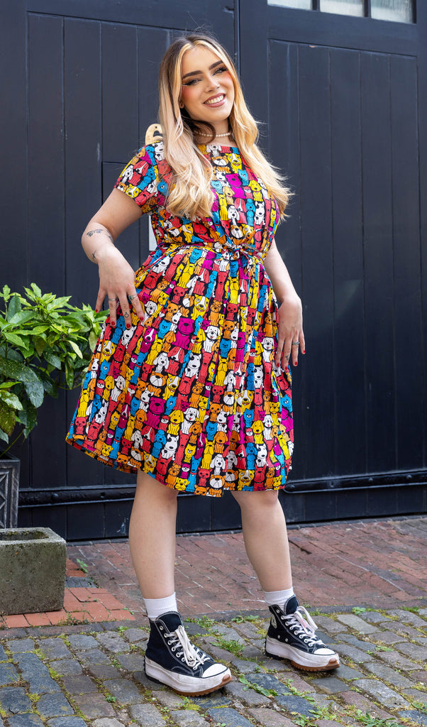 Model with long blonde hair wearing Finding Fox Dog Tea Stretch Dress with Pockets paired with a pearl choker and black trainers with white socks. The dress has a black background with various dog breeds in bright yellow, blue, pink, orange, red and white printed all over. Model is smiling at camera with hands on hips.