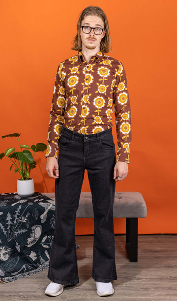 Jack is stood in a studio space wearing the Black Stone Wash Regular Rise Bell Bottom Super Flares with the longsleeve retro brown flowers shirt and white trainers. He has mid length blonde hair and round glasses, he is stood facing the camera with both arms resting by his side. The denim jeans trousers are regular rise with a stonewashed lighter black colour, fitted to thigh and flared bottoms.