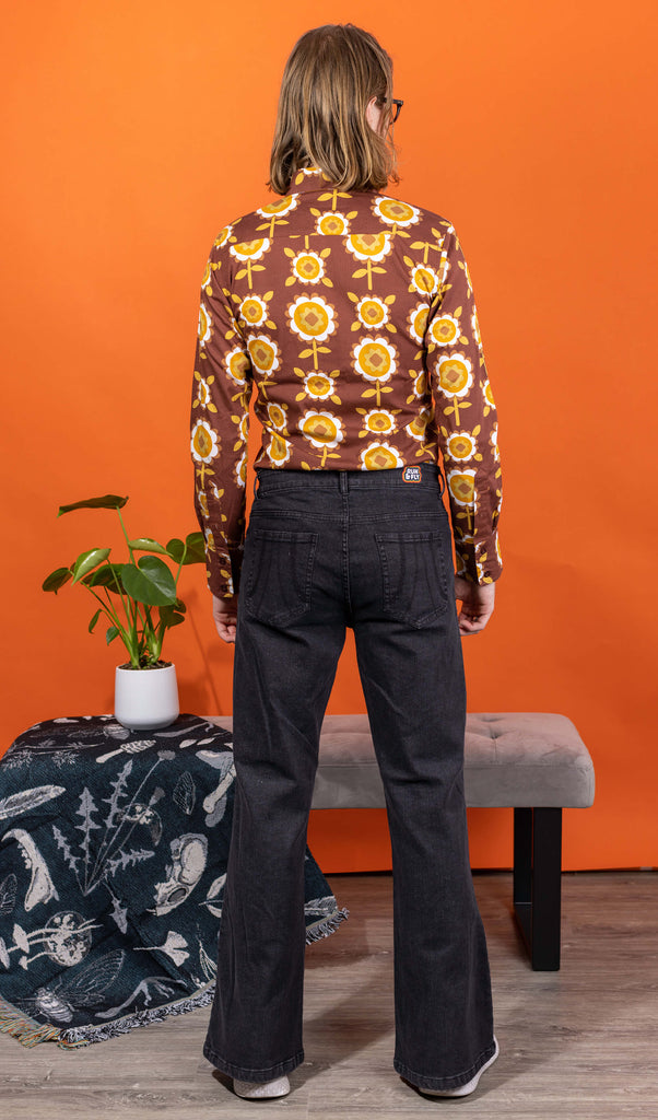 Jack is stood in a studio space wearing the Black Stone Wash Regular Rise Bell Bottom Super Flares with the longsleeve retro brown flowers shirt and white trainers. He has mid length blonde hair and round glasses, he is stood facing away from the camera with both arms resting by his side. The demim jeans trousers are regular rise with a stonewashed lighter black colour, fitted to thigh and flared bottoms.
