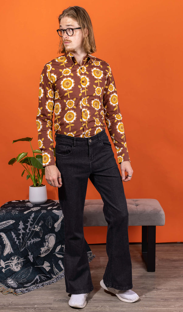 Jack is stood in a studio space wearing the Black Stone Wash Regular Rise Bell Bottom Super Flares with the longsleeve retro brown flowers shirt and white trainers. He has mid length blonde hair and round glasses, he is stood facing the camera looking off to the left with both arms resting by his side. The denim jeans trousers are regular rise with a stonewashed lighter black colour, fitted to thigh and flared bottoms.