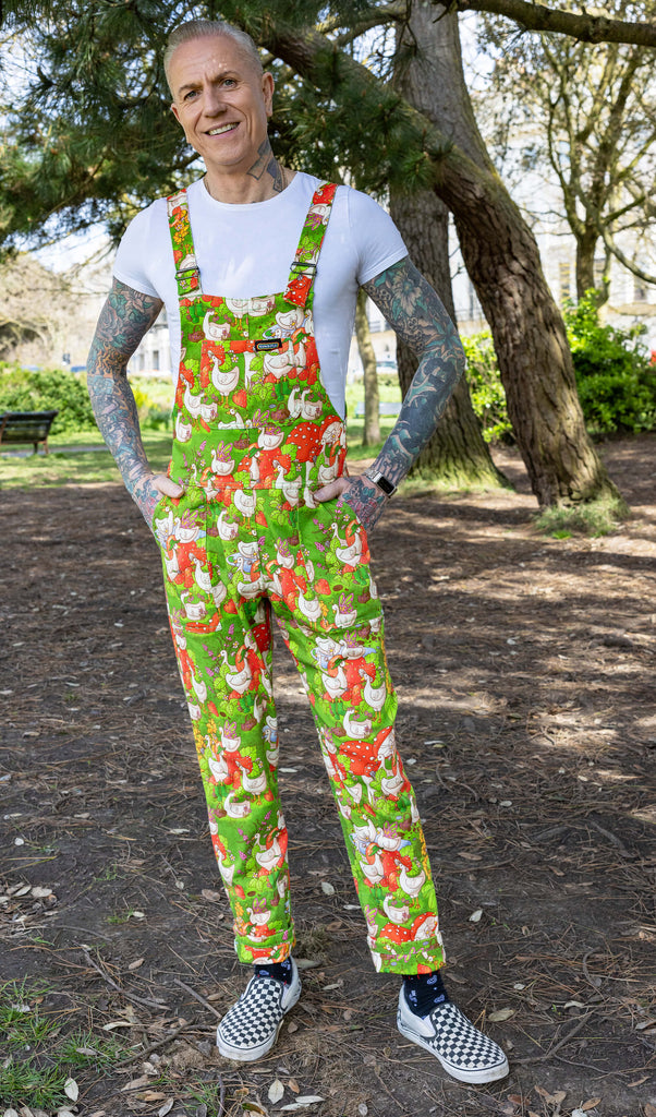 James wearing the Run & Fly x The Mushroom Babes In The Geese Garden Stretch Twill Dungarees with a white tshirt and checkerboard van shoes. James has silvery short hair with lots of tattoos, he is stood in forested park with his arms in the dungaree pockets smiling looking down to camera. The print features geese wearing various cute hats, such as mushrooms and frogs, collecting flowers and strawberries on green grass.