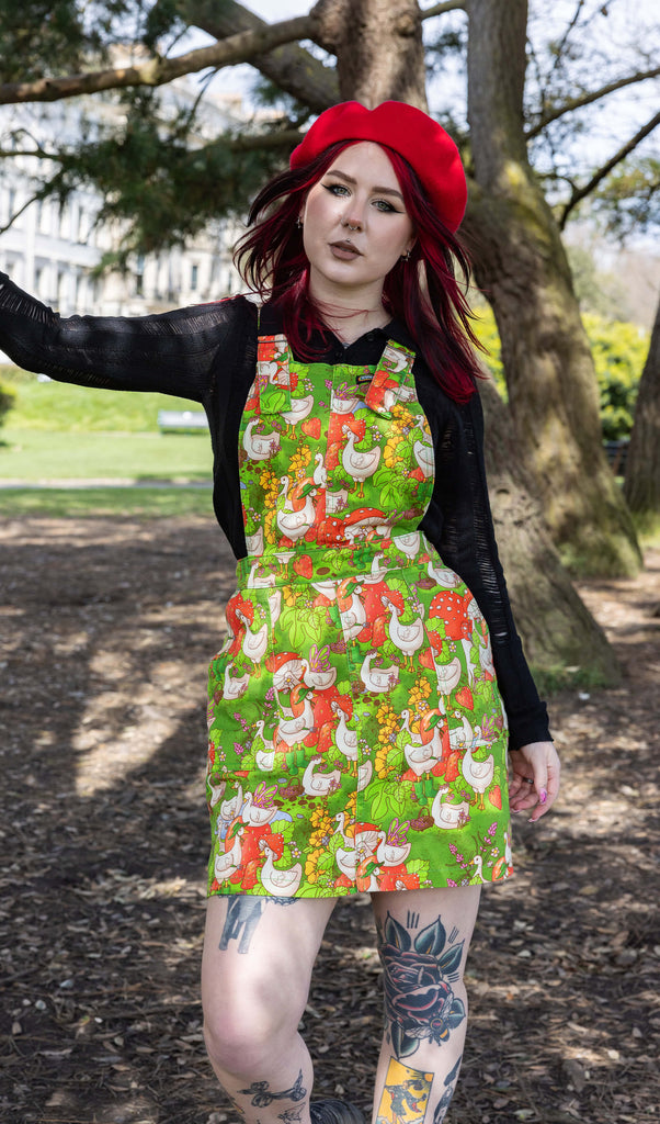 The Run & Fly x The Mushroom Babes In The Geese Garden Stretch Twill Pinafore Dress being worn by Flo outside in a park with a long sleeve black top, black trainers and red mushroom beret. She has red and black hair and tattoos, she is facing forward smising leaning one hand on a tree. The print features geese wearing various cute hats, such as mushrooms and frogs, collecting flowers and strawberries on green grass.