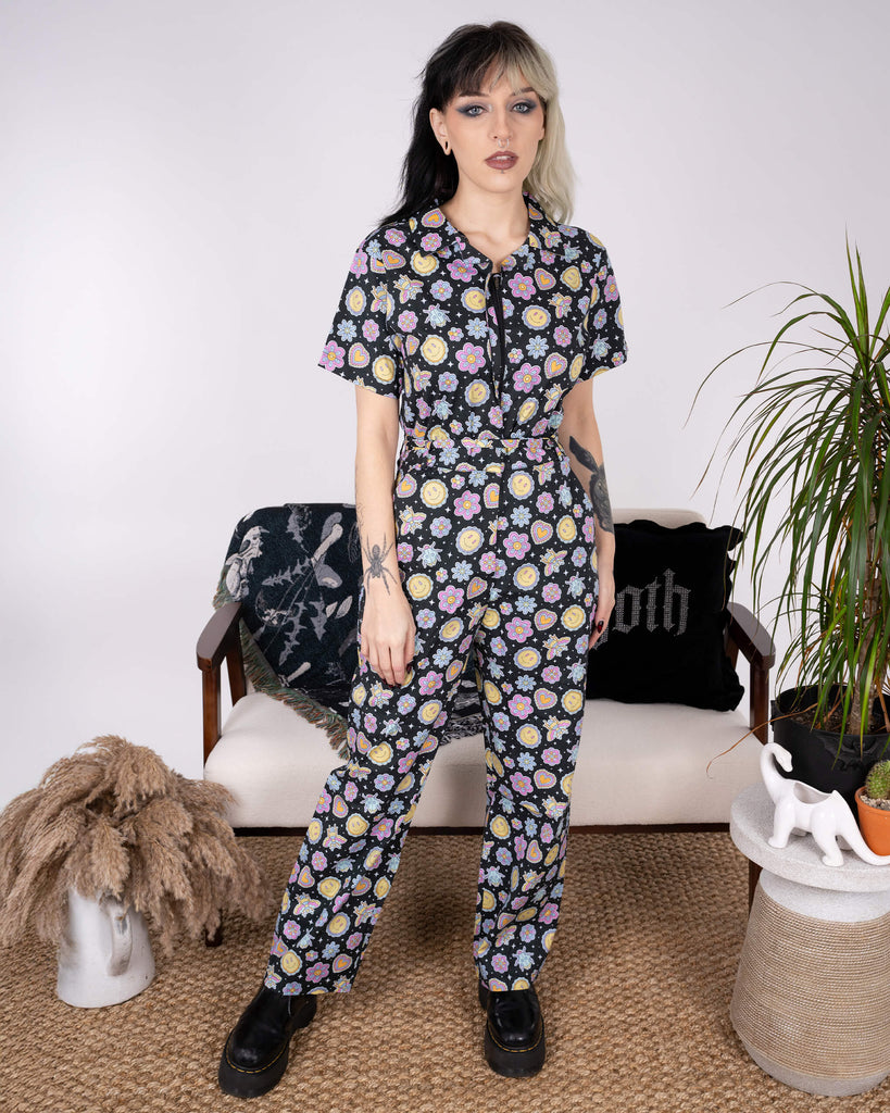 Front view of the Bee Happy Stretch Jumpsuit by Run & Fly. The tailored jumpsuit with its cheerful print of smiley faces, bees, flowers, and hearts is paired with platform boots. A standout ethical slow fashion piece.