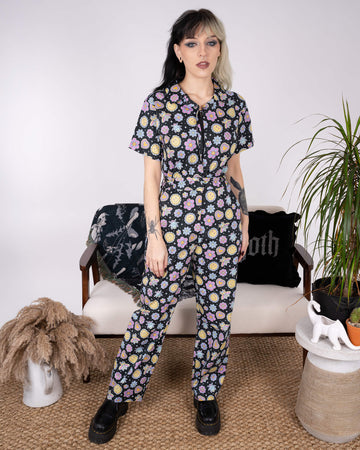 Front view of the Bee Happy Stretch Jumpsuit by Run & Fly. The tailored jumpsuit with its cheerful print of smiley faces, bees, flowers, and hearts is paired with platform boots. A standout ethical slow fashion piece.