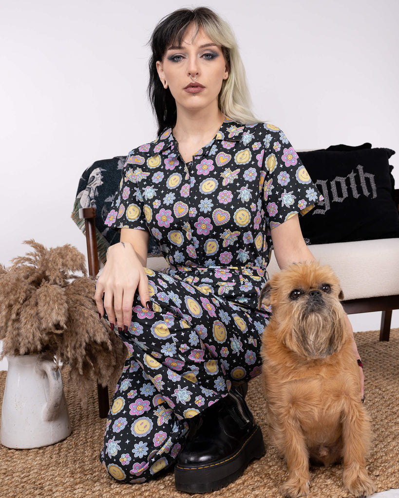 The Bee Happy Stretch Jumpsuit by Run & Fly styled with chunky boots, featuring a cheerful print of flowers, hearts, bees, and smiley faces. The model kneels next to a small dog, showcasing the jumpsuit's comfort and ethical slow fashion credentials.