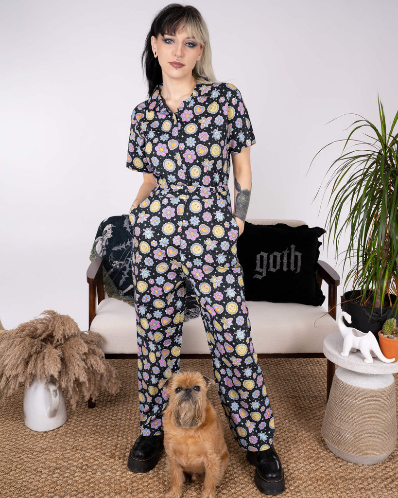 Full-length shot of the Bee Happy Stretch Jumpsuit by Run & Fly, styled with black platform boots. The jumpsuit's fun print of smiley faces, hearts, bees, and flowers makes it a standout piece for ethical slow fashion lovers. A small dog poses adorably with the model.