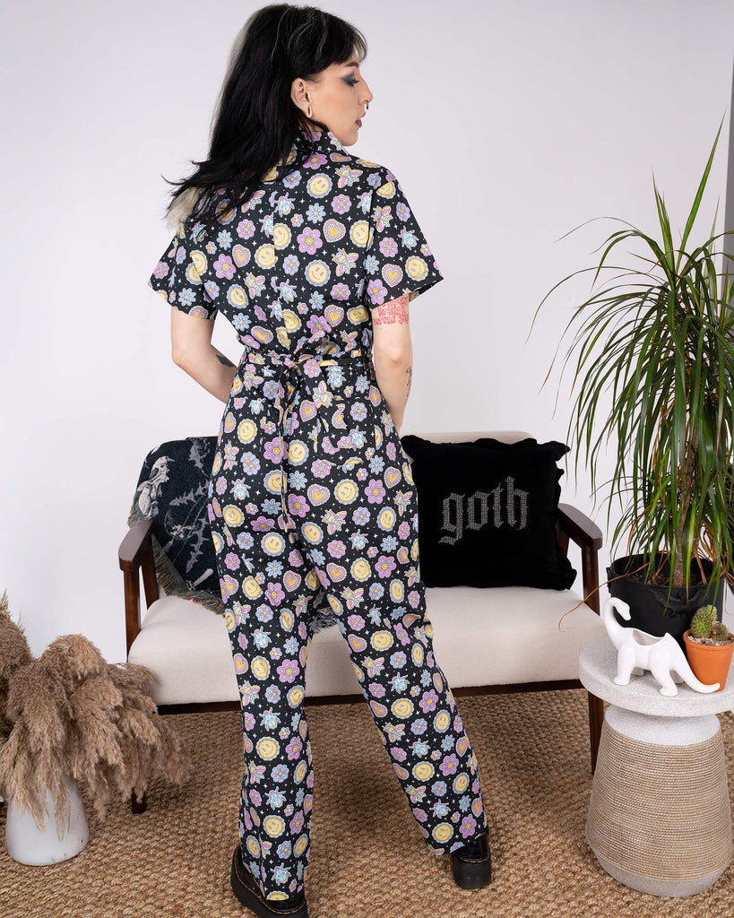 Back view of the Bee Happy Stretch Jumpsuit by Run & Fly, showcasing its tailored fit and tie-back detail. The jumpsuit's playful print of smiley faces, bees, and floral motifs pops against the black base. Perfect for ethical slow fashion lovers.