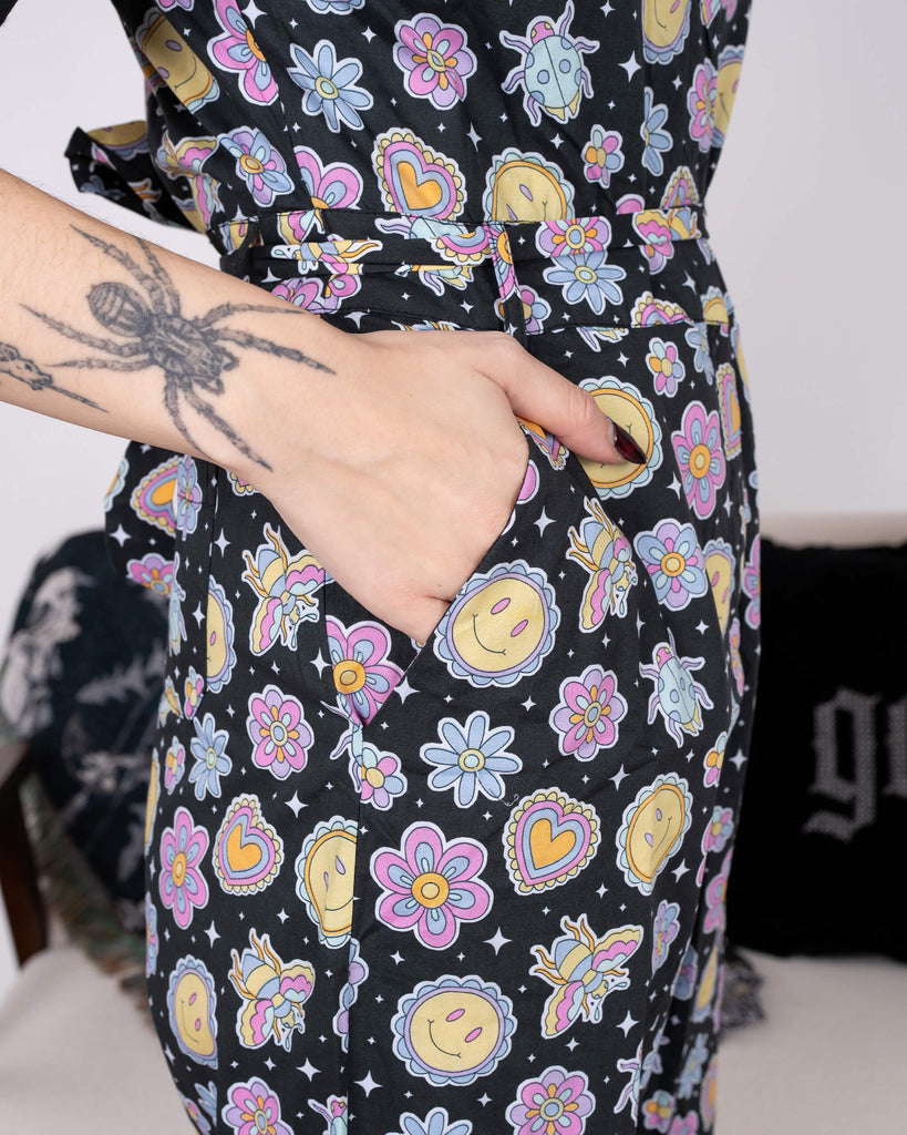Close-up of a hand in the pocket of the Bee Happy Stretch Jumpsuit by Run & Fly. The jumpsuit features a colourful print of cheerful smiley faces, flowers, hearts, and bees on a black background. A gothic-inspired spider tattoo is visible on the model's arm. Ethical slow fashion at its finest.