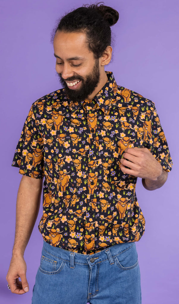 Richard, a hispanic male model with dark hair in a bun and a beard, is stood in a photography studio in Hove in front of a lilac backdrop wearing Highland Cows Short Sleeve Shirt with blue jeans. The black shirt has an all over print of highland cows amongst purple flowers and grass. Richard is facing the camera smiling and looking down to one side with one arm touching the shirt and other down by his side.