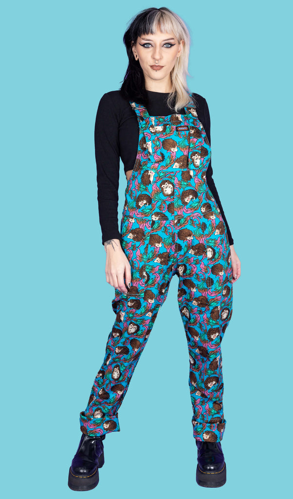 model with black and blonde split dye hair is wearing Hedgehog Stretch Twill Dungarees paired with a long sleeve black top underneath and black boots. The dungarees are blue with cute hedgehog and filigree print. The legs of the dungarees are turned up at the bottoms and the model is stood facing the camera.