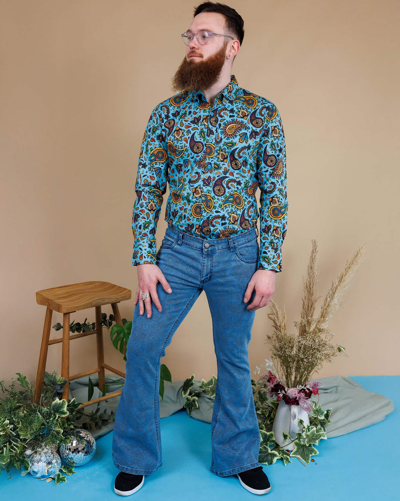 Dec a white male in his 20s with a beard and glasses is stood in a photography studio in Hove wearing New Fit Retro Stretch Stone Wash Denim Hendrix Paisley Bell Bottom Flares with Sky Blue Paisley Short Sleeve Shirt and black shoes. Dec is posing facing the camera with one leg bend and is looking off into the distance.