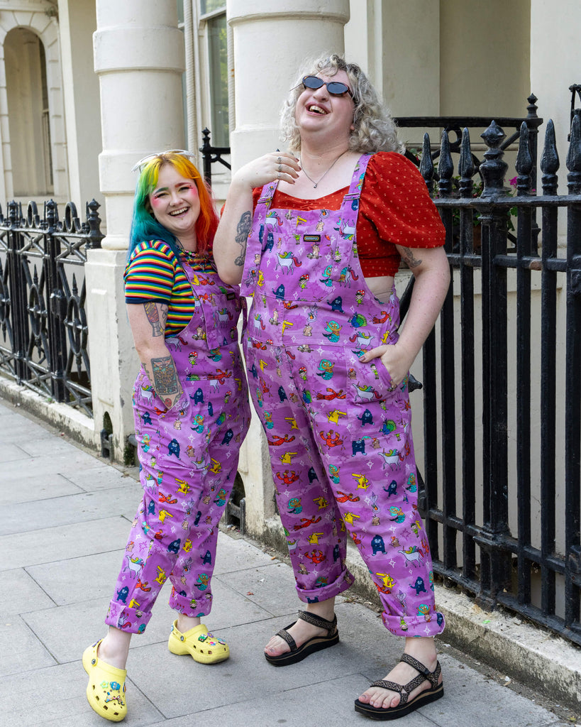 Two models stand side by side, laughing and posing in matching purple dungarees from Run & Fly, covered in playful mythical creatures. One model has rainbow-coloured hair, while the other has blonde curls and wears small sunglasses. Their quirky and colourful outfits radiate fun, with the dungarees paired with casual tops and sandals. Run & Fly is a small, ethical, slow fashion company.