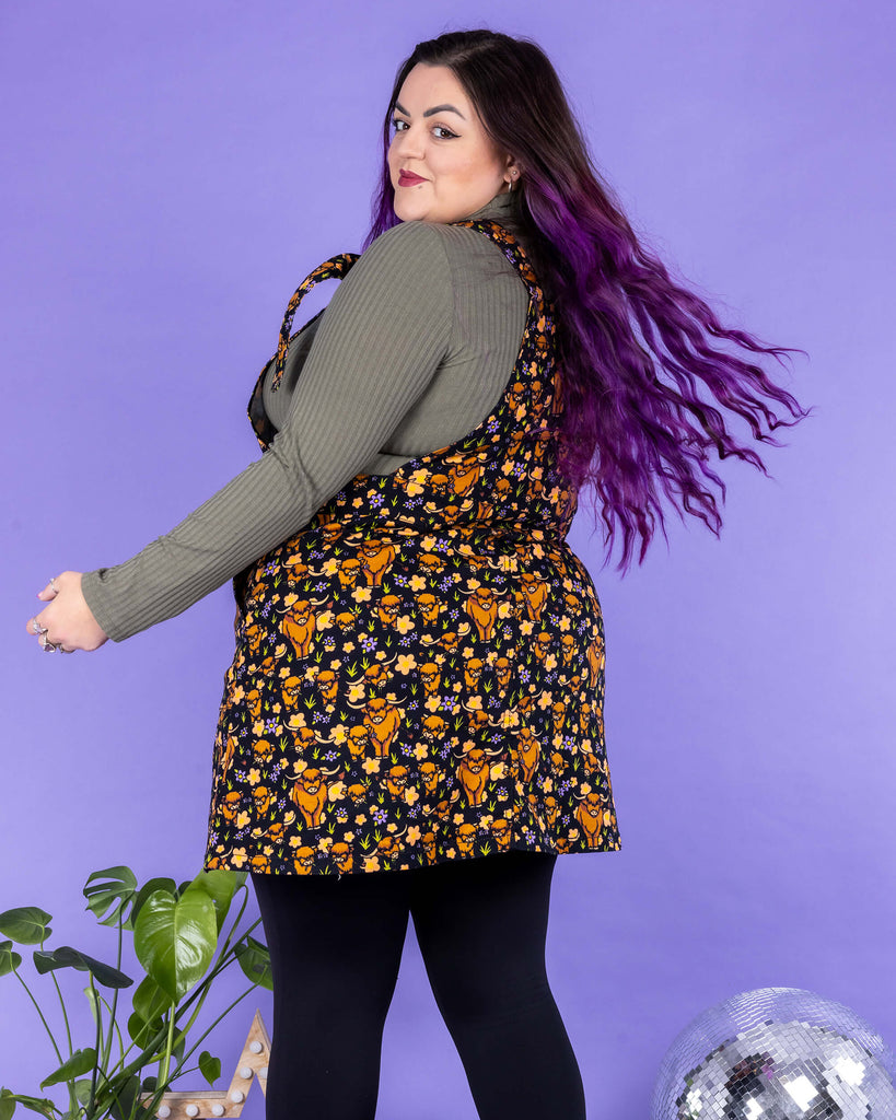 Luisa, a femme model with purple hair, is wearing Highland Cows Stretch Twill Pinafore Dress with a long sleeved khaki turtle neck underneath and black tights and boots. The black pinafore dress has an all over print of Highland cows amongst purple flowers and grass designed by Jen James. Luisa is stood on a black fluffy rug amongst disco balls, a star shaped lamp and a plant in front of a lilac backdrop with their back to the camera and is looking back over their shoulder at the camera.