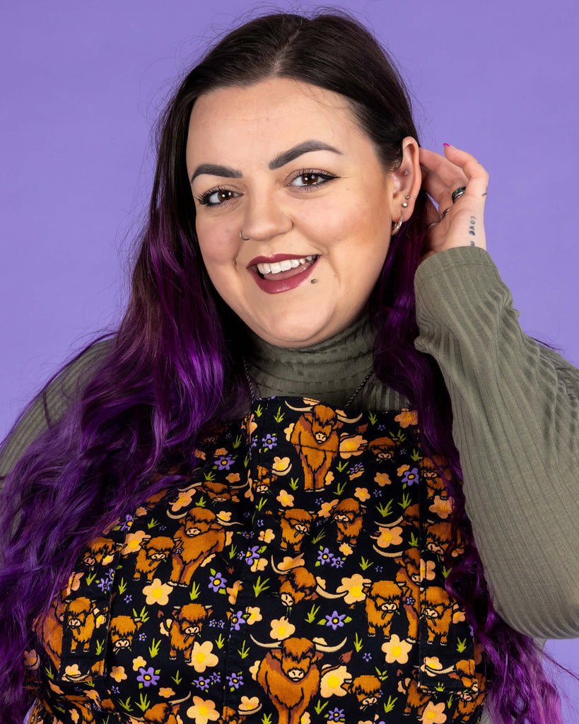 Luisa, a femme model with purple hair, is stood in front of a lilac backdrop wearing Highland Cows Stretch Twill Pinafore Dress with a long sleeved khaki turtle neck underneath. The black pinafore dress has an all over print of Highland cows amongst purple flowers and grass designed by Jen James. Luisa is smiling at the camera and tucking her hair behind her ear. The photo is cropped at the waist.
