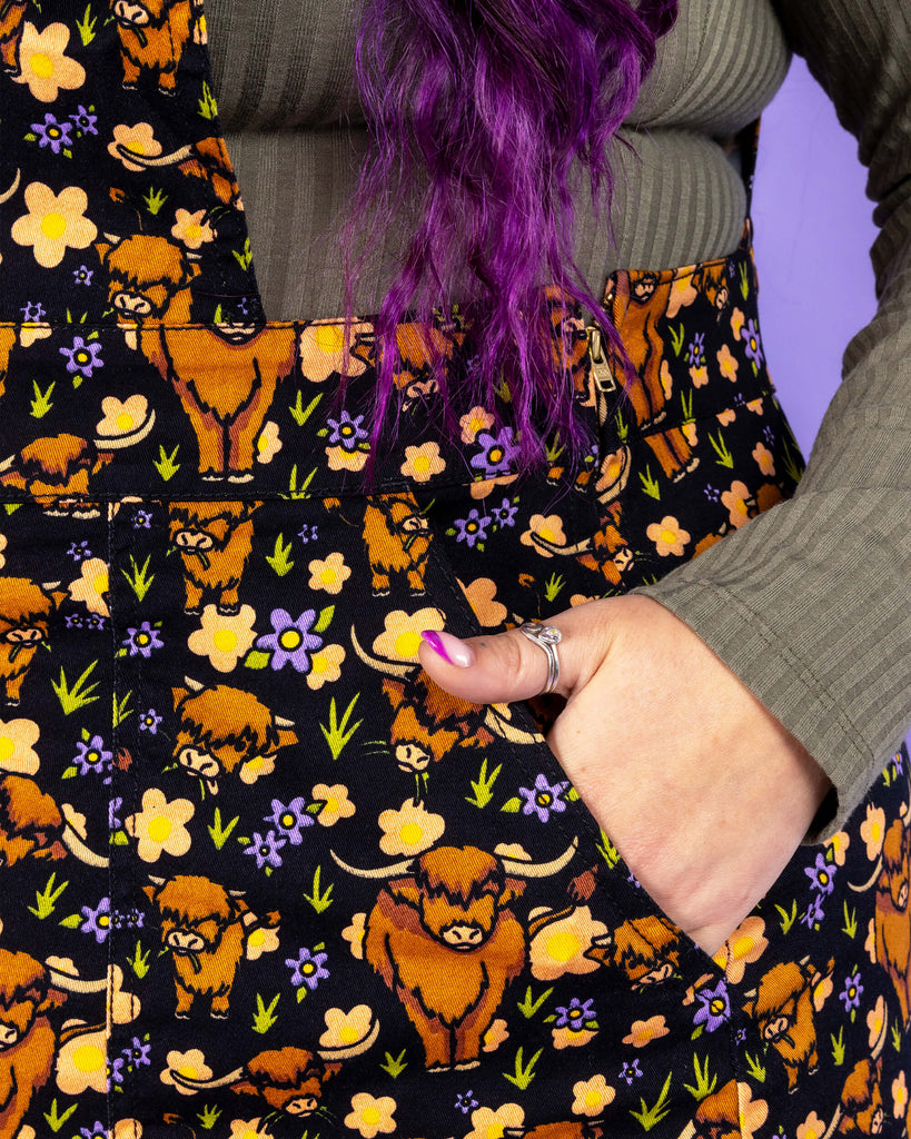 Close up of models hand in the pocket of the Highland Cows Stretch Twill Pinafore Dress. The black pinafore dress has an all over print of Highland cows amongst purple flowers and grass designed by Jen James.