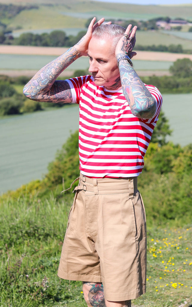 James is wearing the White & Red Stripe Short Sleeve T Shirt with tan coloured shorts, showing off his many tattoos in a grassy field. His hands are pushing his hair back