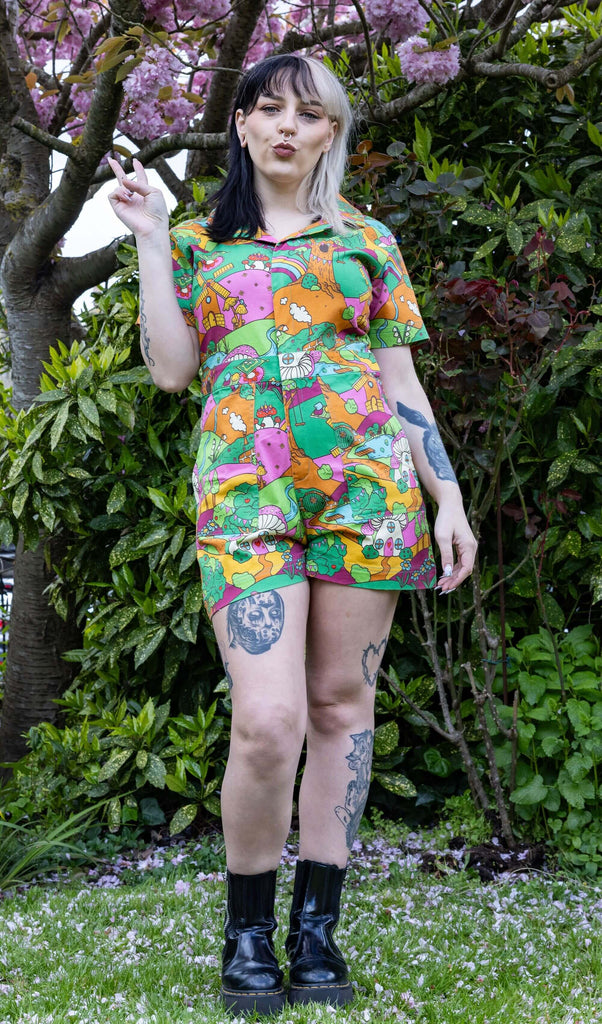 The Run & Fly X The Mushroom Babes Hills Stretch Twill Playsuit worn by a femme tattooed model with split blonde and black hair with black boots. She is stood outside on grass underneath a blossom tree, she is facing forward throwing up a peace sign and pouting. The stretch twill playsuit print features green, pink and orange hills with trees, mushroom cottages, ponds, leading pathways and picnics.
