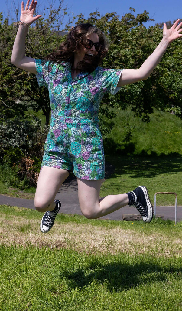 The Succulents Stretch Twill Playsuit worn by a femme model with long brown and blonde hair with black trainers and sunglasses. She is jumping in the air whilst smiling outside in a grassy sunny park area. The playsuit print features various sized succulents in pastel blue, pastel lilac, teal, pink and pale yellow.