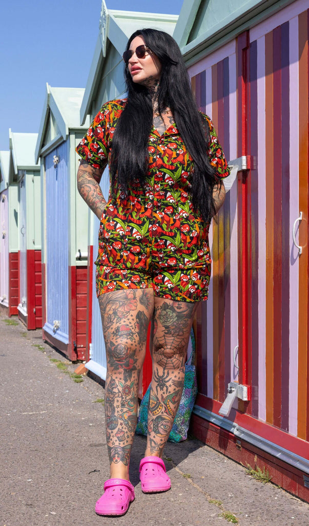 The Red Panda Stretch Twill Playsuit modelled by a femme tattooed model with long black hair with pink croc shoes and black sunglasses. She is outside along the beach with colourful beach huts behind her, she is facing forward looking off to the left with both hands in the playsuit pockets. The playsuit print is a black base covered with illustrated bamboo, branches and red pandas shown asleep, curled up, hanging off branches and eating the leaves.
