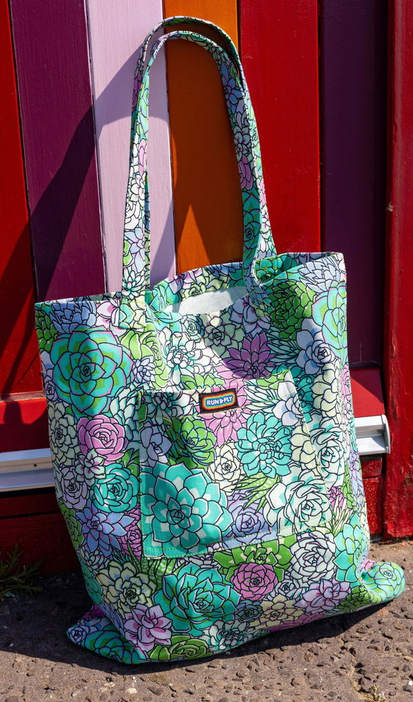 The Succulents Tote Bag leaning up against a colourful beach hut along hove seafront. The tote bag has a front pocket with a rainbow run and fly label in the top middle. The succulent print features pastel blue, pastel teal, green, purple and pale yellow toned succulent plants with black outlines.