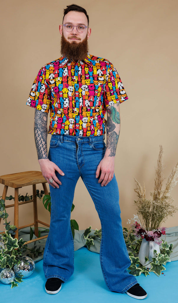 Declan a man in his 20's with a beard, tattoos and glasses is wearing a pair of blue denim flares with a shirt with all over dog print on and is standing against a beige background with a stool, florals and mirrorball