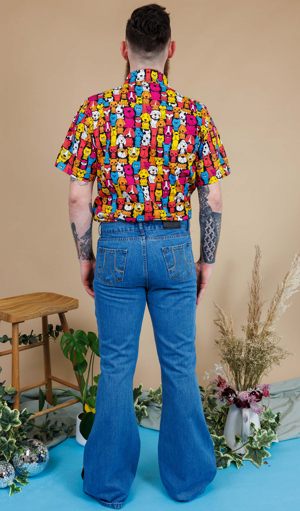 Back shot of Declan a man in his 20's with a beard, tattoos and glasses is wearing a pair of blue denim flares with a shirt with all over dog print on and is standing against a beige background with a stool, florals and mirrorball
