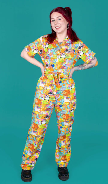 Florence a red haired female model with tattoos and cool make up is wearing a colourful rainbow jumpsuit with cute drawing by Katie Abey. She is standing against a turquoise background and smiling at camera. 