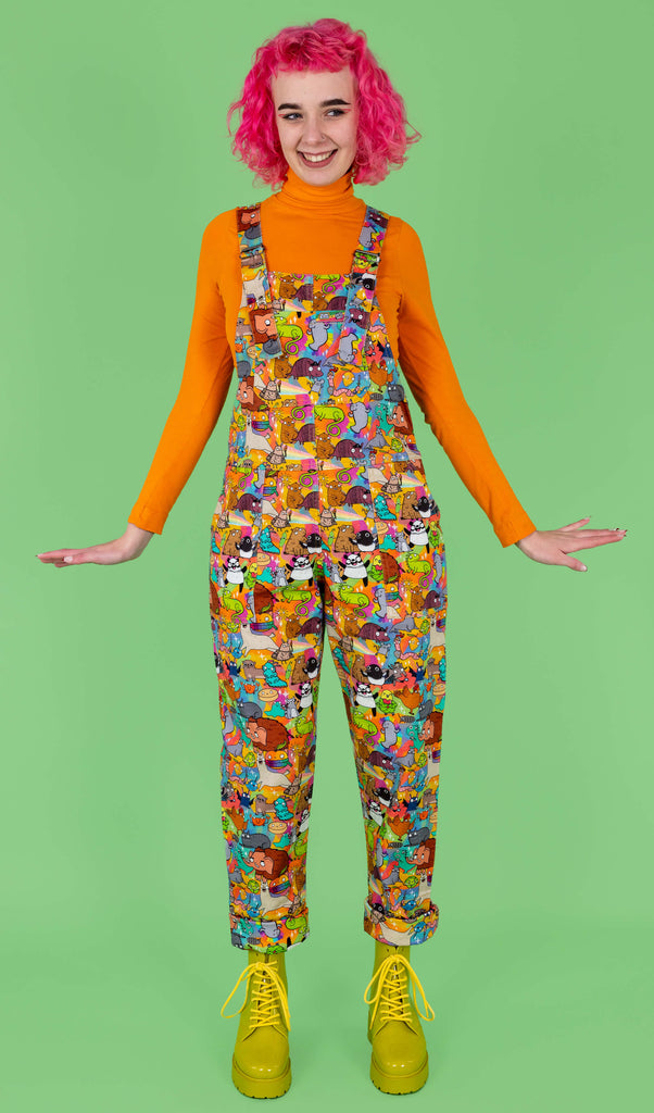 Lottie a pink hair tall female white model is standing with her arms out wearing a pair of dungarees with characters by Katie Abey all over them. She is smiling and looking away from camera. She has an orange roll neck and green boots on and is against a light green background.