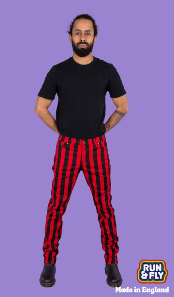 Richard, a hispanic male model with dark hair in a bun and a beard, is stood in a photography studio in Hove in front of a lilac backdrop wearing Black & Red 1" Striped Mid Rise Stretch Skinny Jeans with a black t shirt and black shoes. Richard is smiling and facing the camera with his arms behind his back and legs slightly apart. Run & Fly logo is at the corner or the photo with 'Made in England' written underneath.