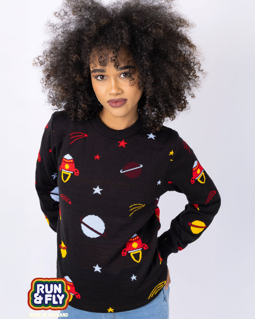 Cindy is stood in front of a white studio background wearing the retro rocket outer space jumper with light denim jeans. They are facing forward posing with both hands on their hips and smiling to camera. Photo is cropped from the hips up.