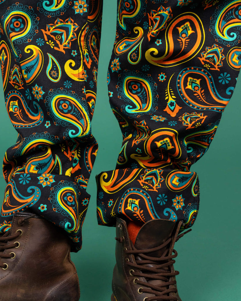 Detailed close-up of Run & Fly dungarees' leg area, showcasing the intricate paisley pattern in bright orange, teal, and yellow against a black background. The model pairs the dungarees with brown boots, emphasising the bold and ethical fashion statement.