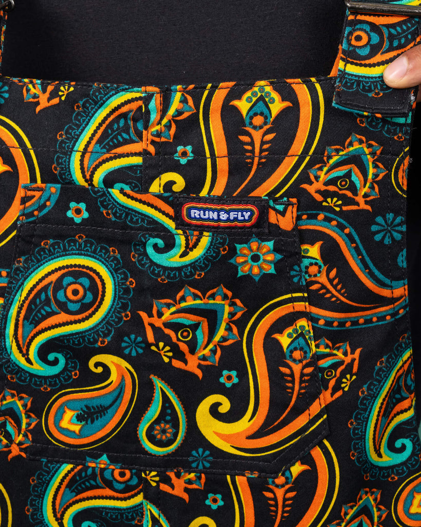 Close-up view of the chest area of Run & Fly dungarees with a vibrant paisley pattern in orange, teal, and yellow on a black background. The model wears a black t-shirt underneath, highlighting the detailed design and the brand's commitment to ethical slow fashion.