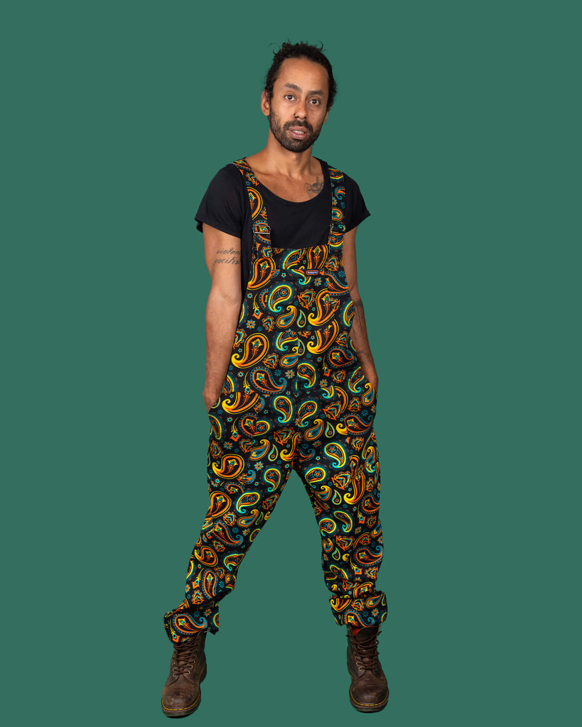 Model wearing Run & Fly dungarees with a bold paisley pattern in orange, teal, and yellow on a black background. The dungarees are styled with a black t-shirt and brown boots, embodying the brand's ethical and slow fashion principles.