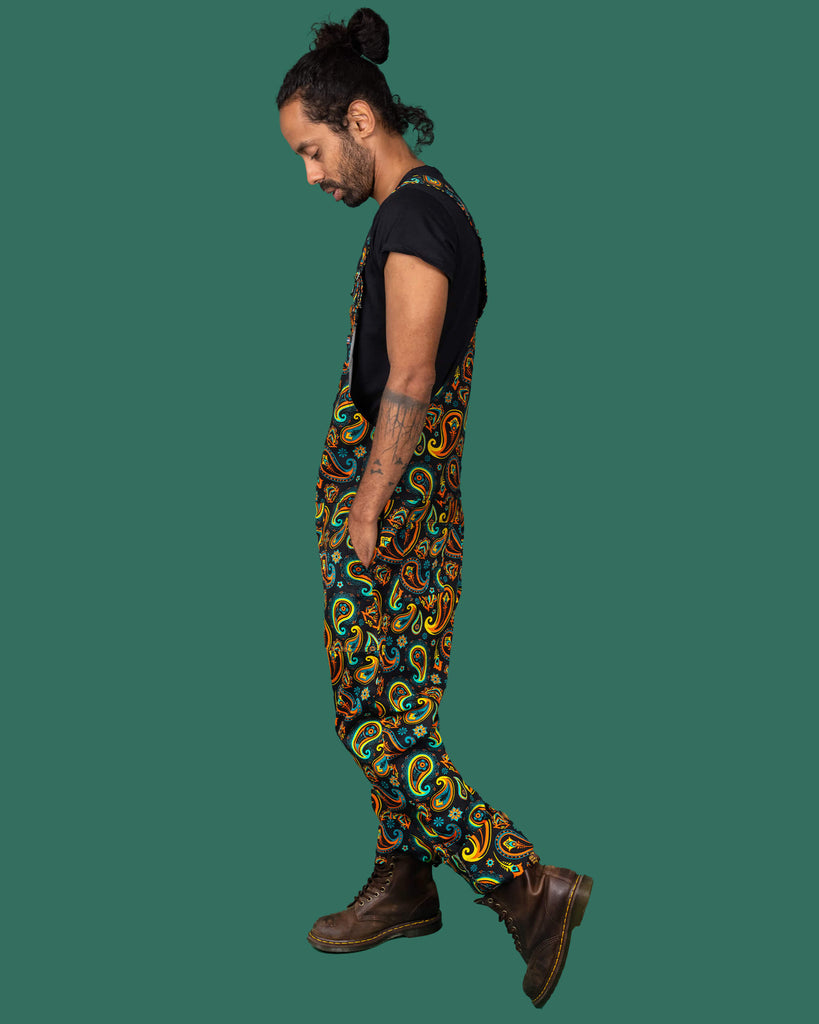 Side view of a person wearing Run & Fly's colourful paisley patterned dungarees, paired with a black t-shirt and brown boots. The side pose highlights the dungarees' comfortable fit and the vibrant design.