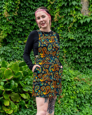 Model wearing a Run & Fly ethical slow fashion black pinafore dress with a vibrant paisley print, paired with a black long-sleeve top, chunky platform boots, and visible leg tattoos, posing outdoors against a backdrop of lush green ivy.