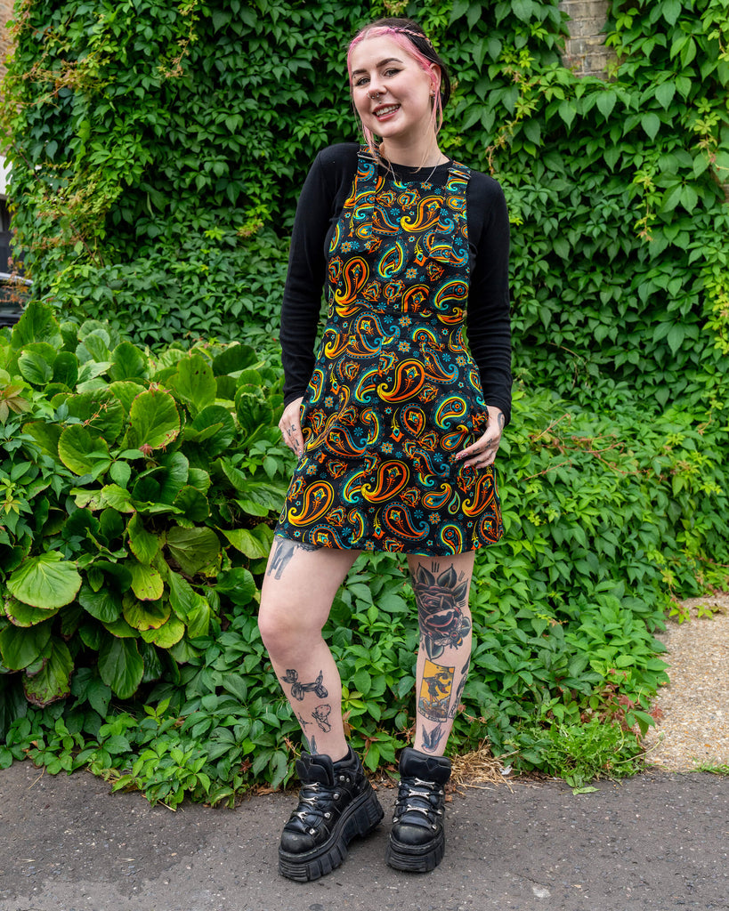 Model wearing a Run & Fly ethical slow fashion black pinafore dress with a vibrant paisley print, paired with a black long-sleeve top, chunky platform boots, and visible leg tattoos, posing outdoors against a backdrop of lush green ivy.