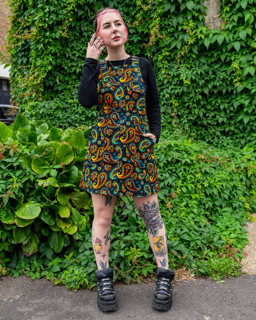 Model wearing a Run & Fly ethical slow fashion black pinafore dress with a vibrant paisley print, paired with a black long-sleeve top, chunky platform boots, and visible leg tattoos, posing outdoors against a backdrop of lush green ivy.